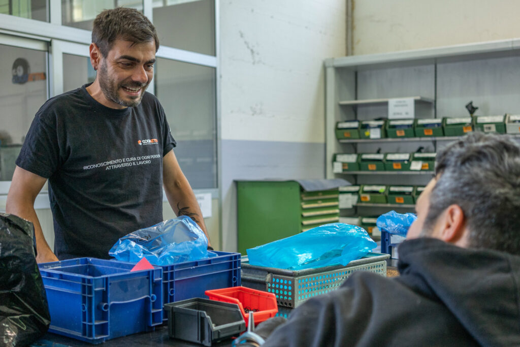 Operatore coopattiva con maglia coopattiva mentre spiega come effettuare una lavorazione nel laboratorio socio occupazionale di frignano. L'uomo e sorridente e ha davanti delle casse di colore blu.