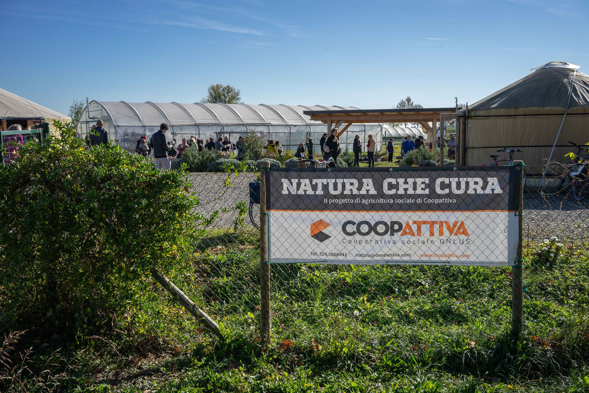 Vista del cartello del campo natura che cura di coopattiva. Dietro si vede il campo con persone che lavorano.