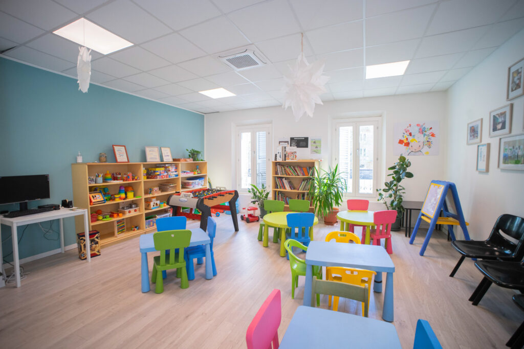 Veduta di un'ambiente degli spazi di mediazione familiare di coopattiva. Sono presenti un biliardino e alcuni tavoli e sedie di plastica altezza bambino. Sulle pareti ci sono libreria e scaffali per giocattoli.