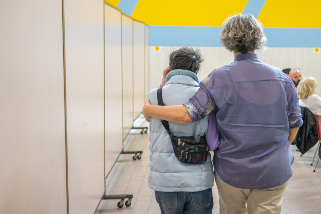 Due donne in ambiente protetto coopattiva, sulla sinistra un'anziana viene accompagnata in modo accogliente da un addetto coopattiva sulla destra.